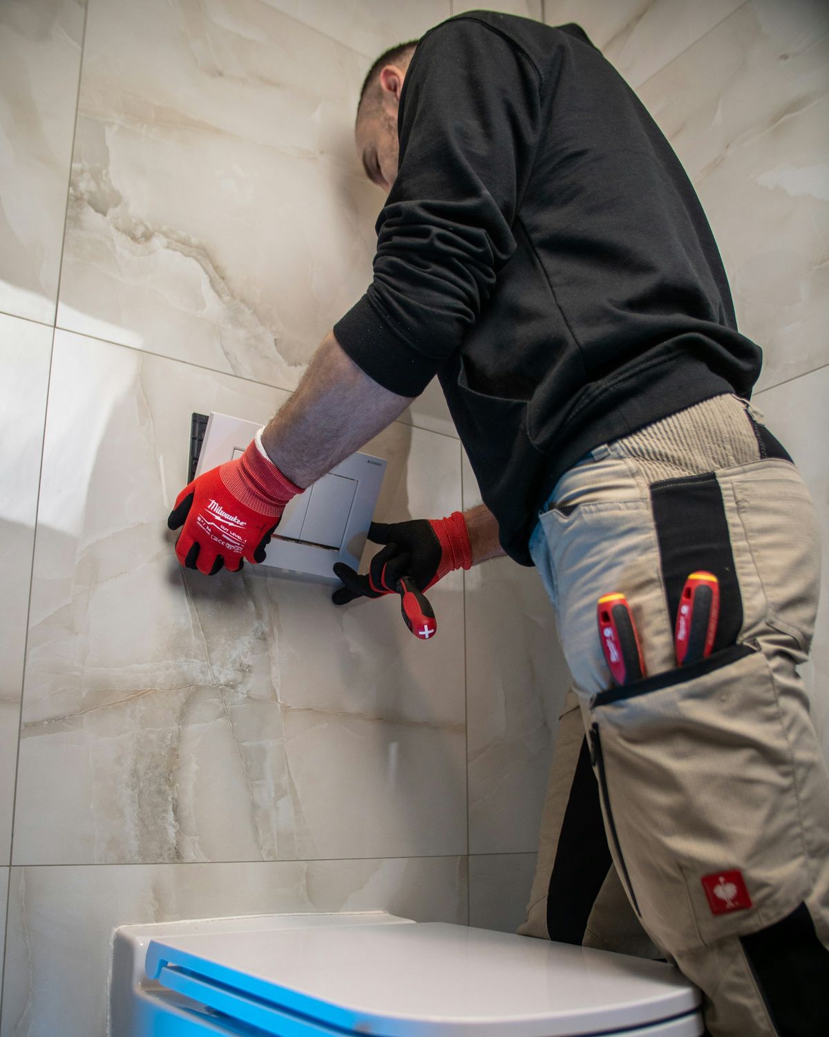 Un plombier, gants aux mains, installe une plaque de commande au mur d'une salle de bain.